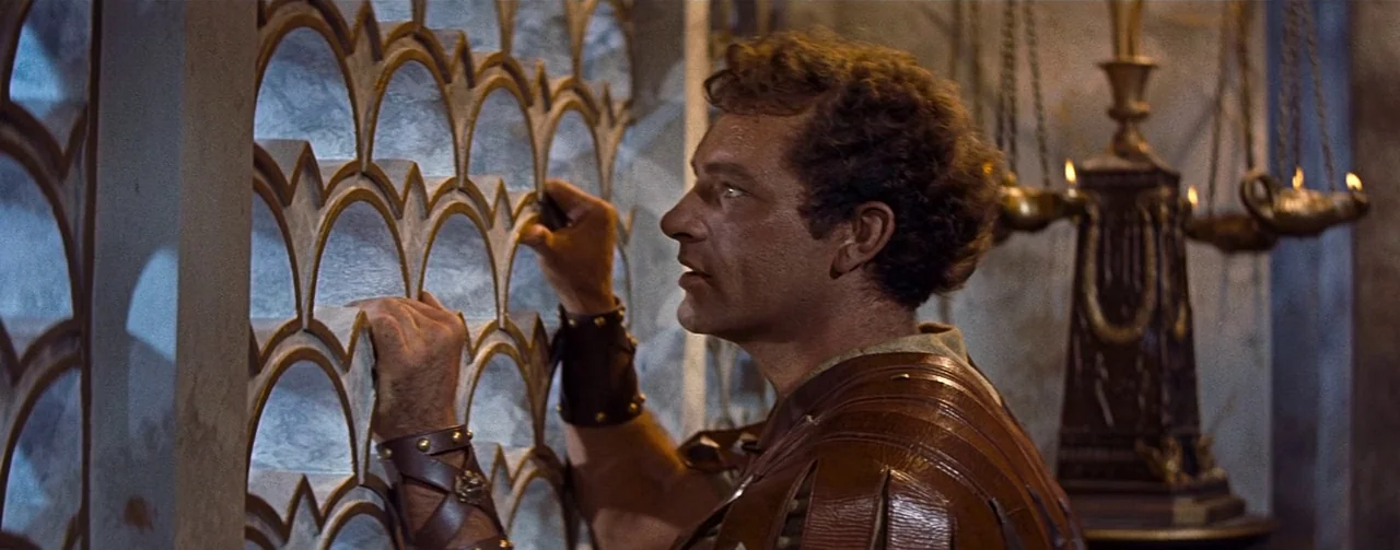 The Robe (1953) movie still: medium — A man wearing ancient Roman-style leather armor peers through a curved metal latticework screen. An ornate oil…