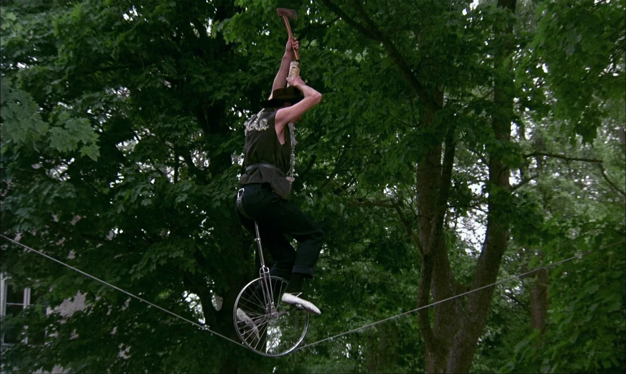 My Life As A Dog (1985) movie still: fullbody — A man balances on a unicycle on a tightrope surrounded by lush green trees. He holds an axe…