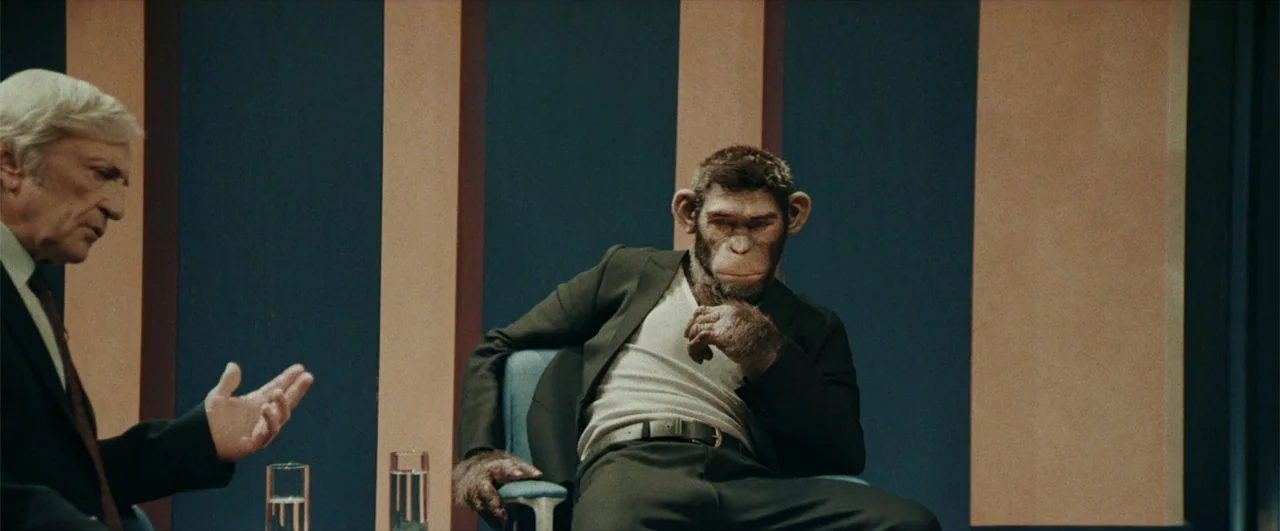 Better Man (2024) movie still: medium — A television studio features vertical blue and tan panels in the background. A man speaks to a chimpanzee…