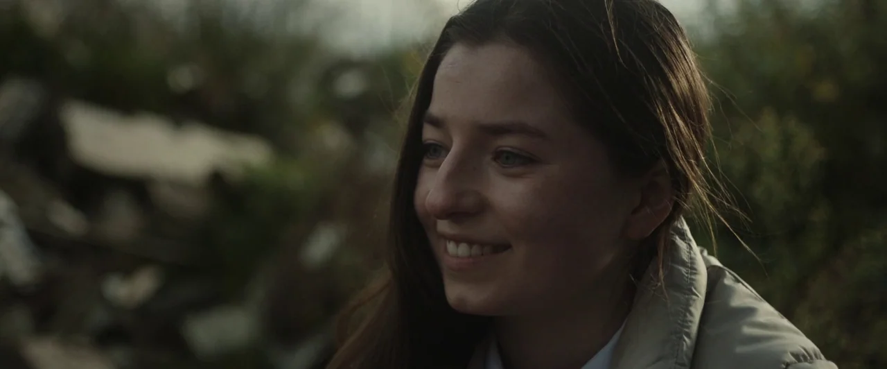 You Are Not My Mother (2021) movie still: closeup — An open grassy field with blurred vegetation visible in the background. A young woman with long…