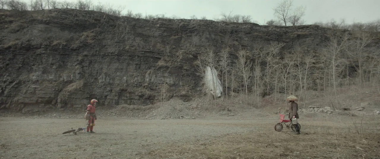 Turbo Kid (2015) movie still: wide — A vast, barren gravel quarry sits beneath a towering, jagged gray rock cliff. Two distinct figures stand facing…
