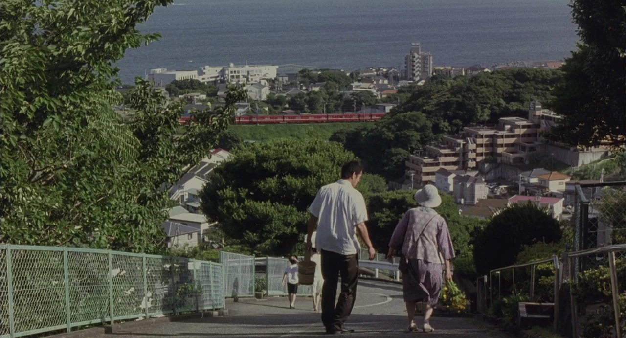 Still Walking (2008) movie still: wide — A sloped residential street descends toward a coastal town with a red train passing. A man and an older…