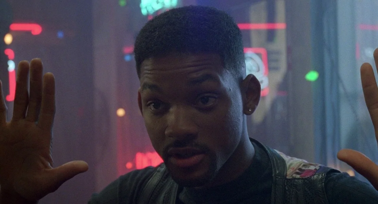 Bad Boys (1995) movie still: closeup — A man stands behind a glass surface against a blurred background of neon signs. He is holding his hands up…