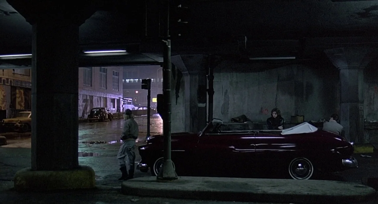 Streets of Fire (1984) movie still: wide — A dark urban environment beneath a concrete bridge support pillar at night. Three individuals stand near…