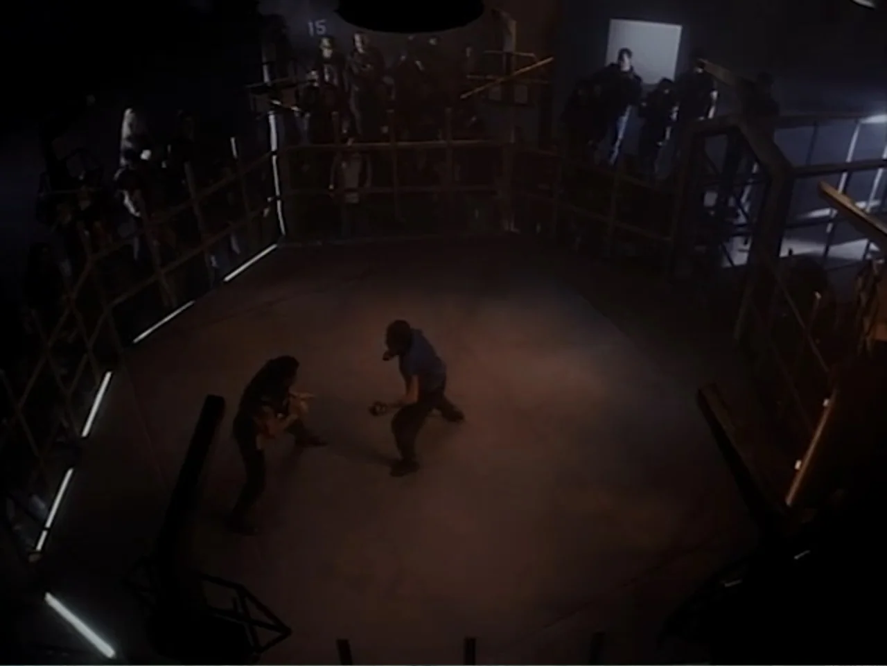 Firepower (1979) movie still: wide — A dimly lit indoor fighting cage is surrounded by wooden railings and an audience. Two athletic men face each…