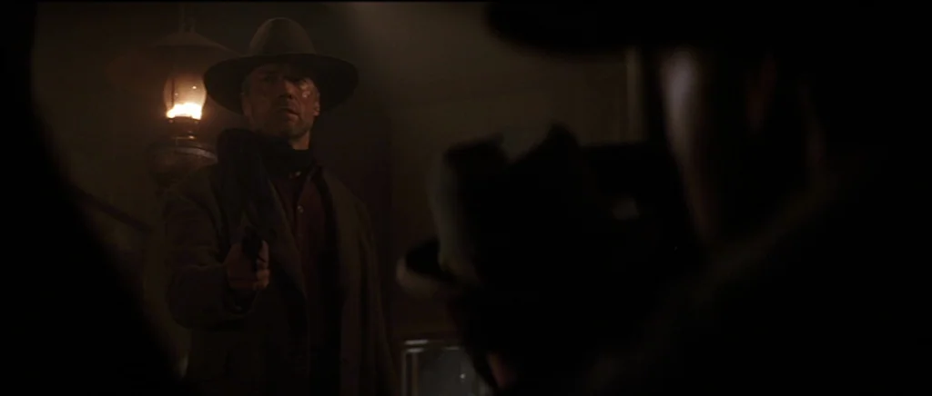 Unforgiven (1992) movie still: medium — A dark, dimly lit 19th-century room features a man holding a handgun towards camera. The man stands near a…