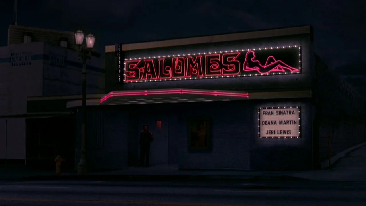 Auto Focus movie still: wide — A dark city street features a nightclub building with prominent pink neon signage overhead. A marquee on the wall…