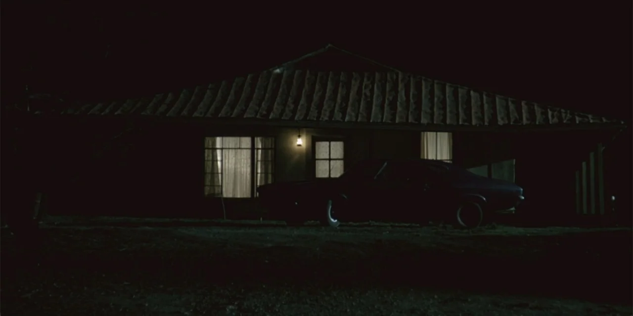 El sótano (2024) movie still: wide — A single-story 1970s house sits in total darkness at night. A dark muscle car is parked in the dirt driveway…