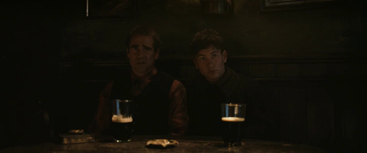 The Banshees of Inisherin (2022) movie still: medium — A dimly lit room with dark wood paneling and shadowed corners. Two men sit together at a…