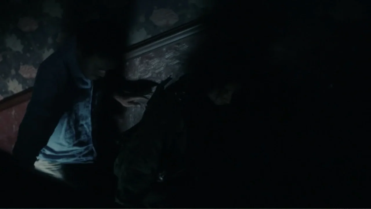 Bel Canto (2018) movie still: medium — A dimly lit indoor stairwell features dark floral wallpaper and a wooden banister. Two individuals move…