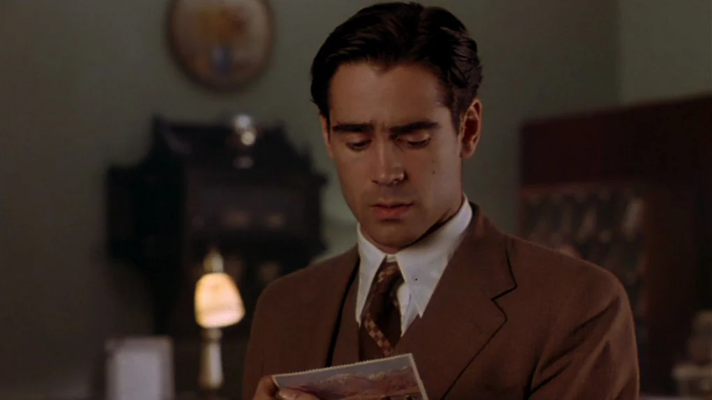 Ask The Dust (2006) movie still: medium — A dimly lit room with a vintage lamp and a framed item on the wall. A man in a brown suit looks down at a…