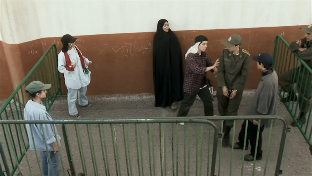 Offside (2015) movie still: wide — An outdoor area is enclosed by green metal barriers against a plain white wall. A diverse group of people stand…