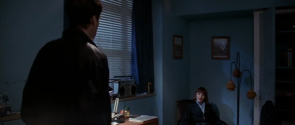 The Crying Game (1992) movie still: medium — A blue-walled room is furnished with a floor lamp and a desk by a window. A woman sits in a chair while…