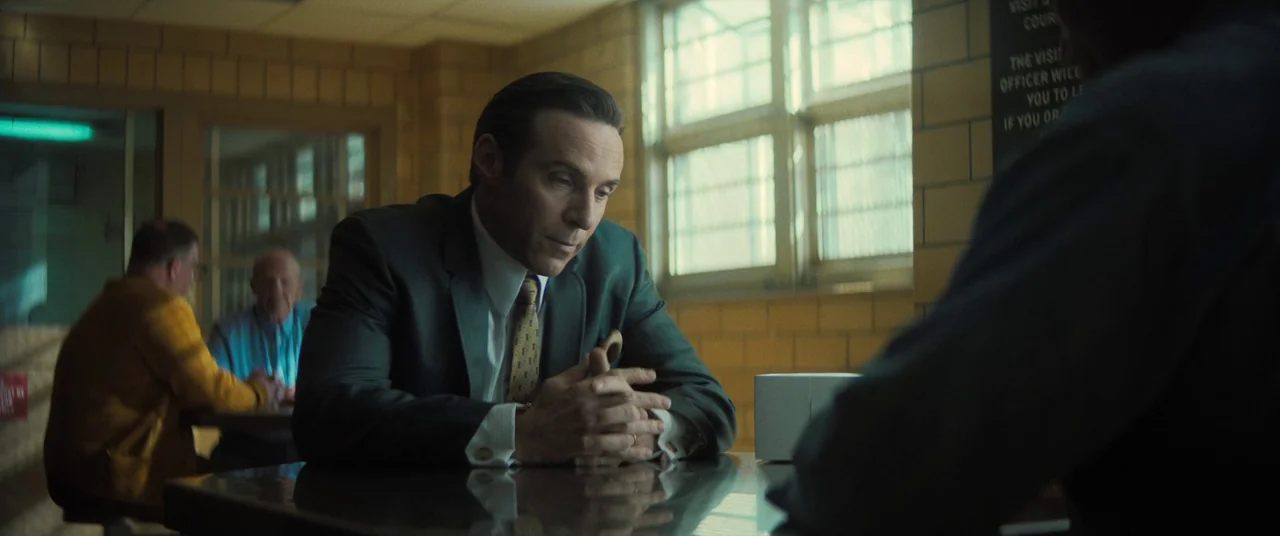 The Many Saints of Newark (2021) movie still: medium — A visitation room has tan brick walls and a metal table surface reflecting light. A man in a…