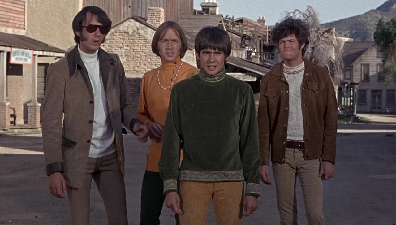 Head (1968) movie still: fullbody — Four men stand on a dirt road in a quiet western town set. Wooden buildings and rustic facades line the…