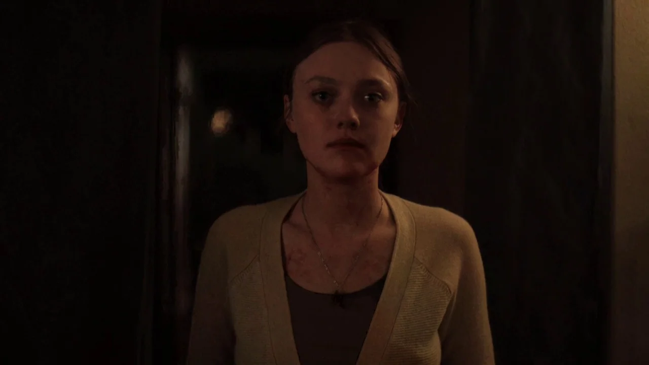 Night Moves (2013) movie still: medium — A dimly lit indoor corridor with dark walls and shadows filling the background space. A young woman stands…