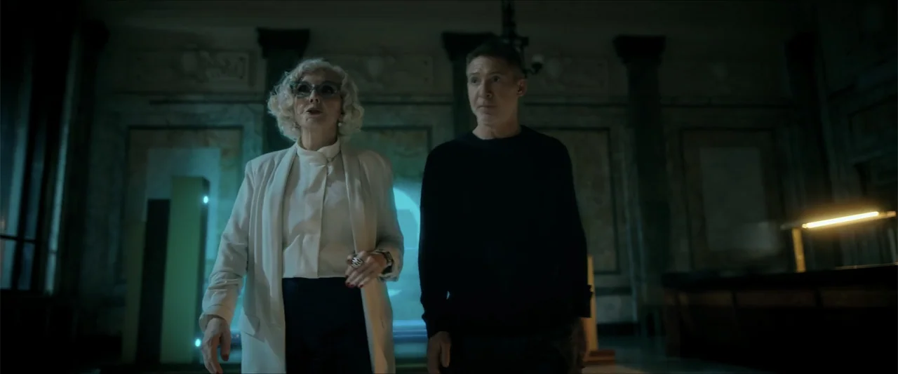 Checkmate (2015) movie still: medium — A dimly lit interior space with classical architectural details and dark marble-like pillars. An older woman…