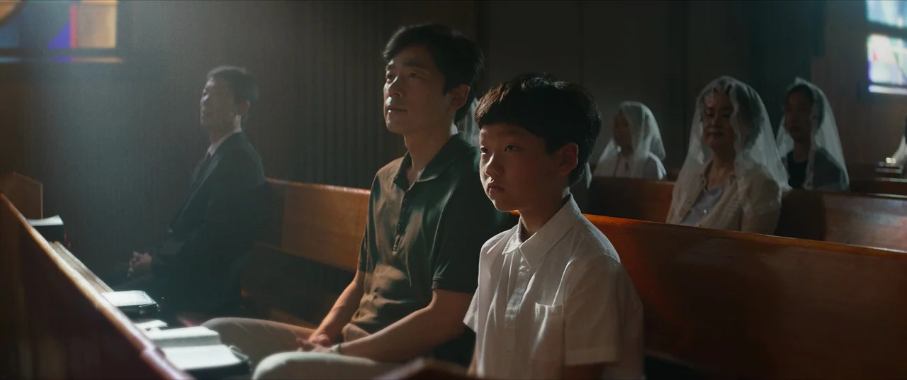 The Divine Fury (2019) movie still: medium — A congregation sits in wooden pews within a dimly lit building with stained glass. A man and a young…