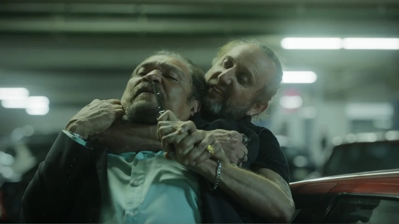 Lecciones para canallas (2022) movie still: medium — A dimly lit concrete parking structure with blurred vehicle forms in the background. A man…