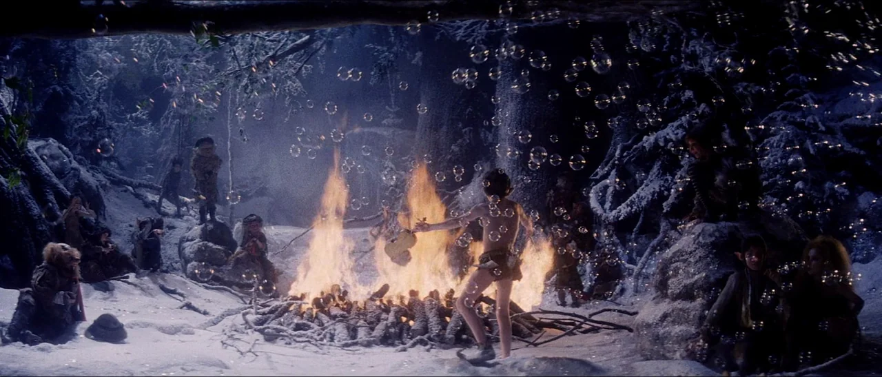 Legend (1985) movie still: wide — A snowy forest clearing features a large burning pile of wooden logs. A small child stands near the fire while…