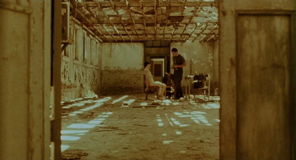 Traffic (2000) movie still: wide — A desolate room with an exposed wooden roof joist structure and peeling plaster walls. One individual sits in a…
