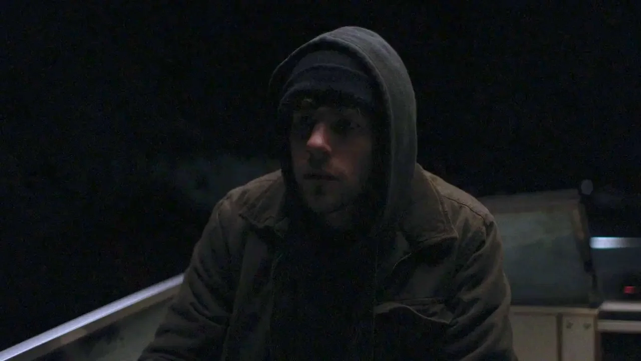Night Moves (2013) movie still: medium — A dark, nighttime environment aboard a small vessel on open water. A man in a hoodie and brown jacket sits…