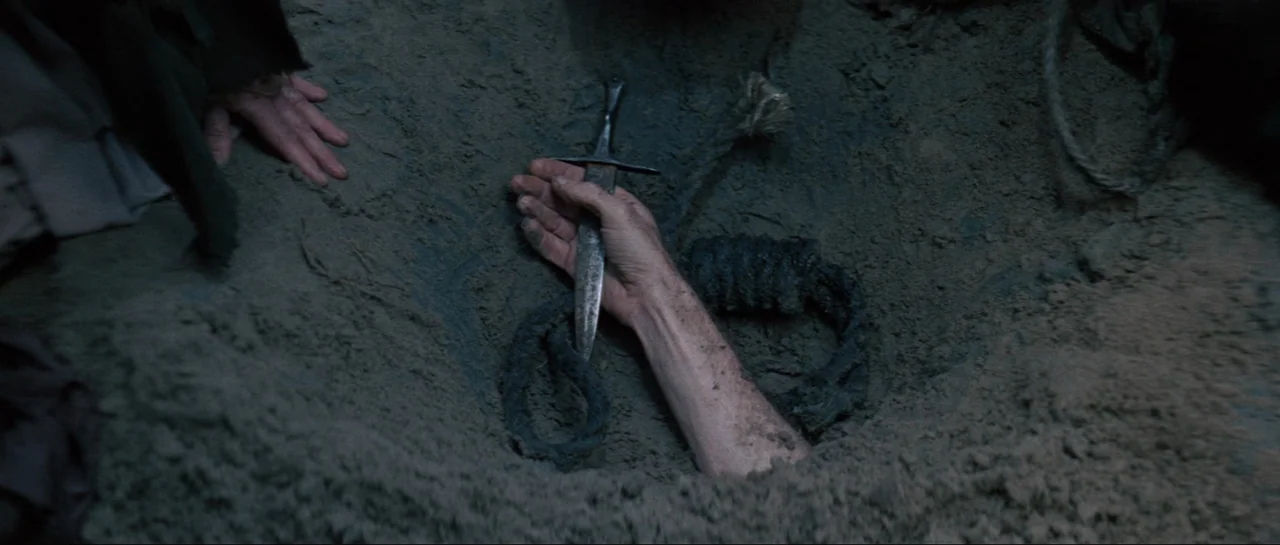Macbeth (1971) movie still: closeup — A shallow hole in the ground is filled with loose, greyish-brown sand. A single human hand holds a small steel…