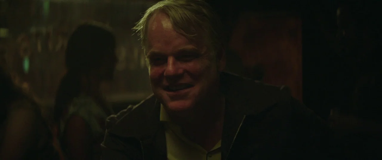 God’s Pocket (2014) movie still: closeup — A dimly lit, low-contrast bar interior with silhouettes of people in the blurred background. A…