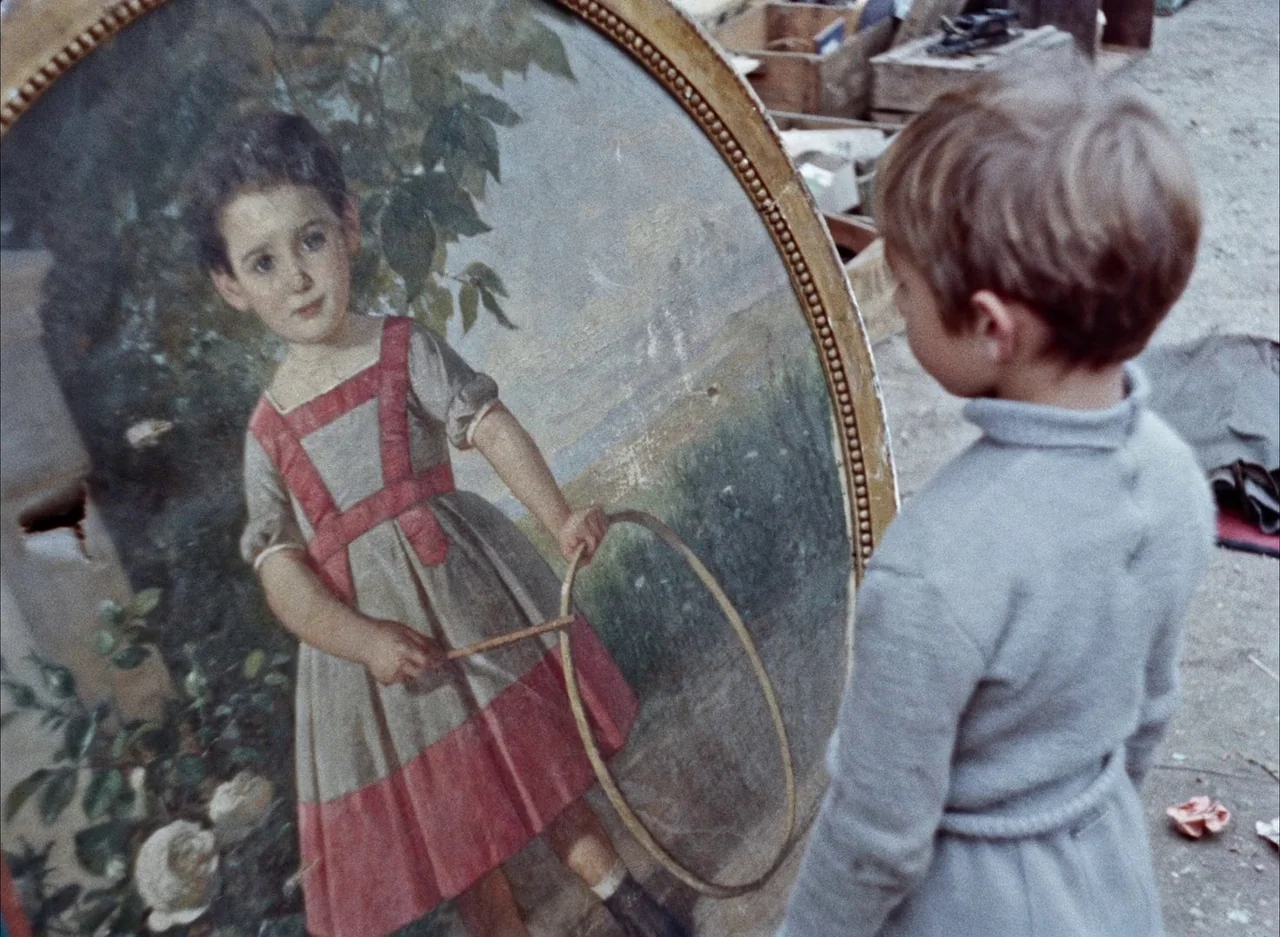 The Red Balloon (1956) movie still: medium — A young boy stands in a cluttered space filled with wooden crates and debris. He gazes at a large oval…