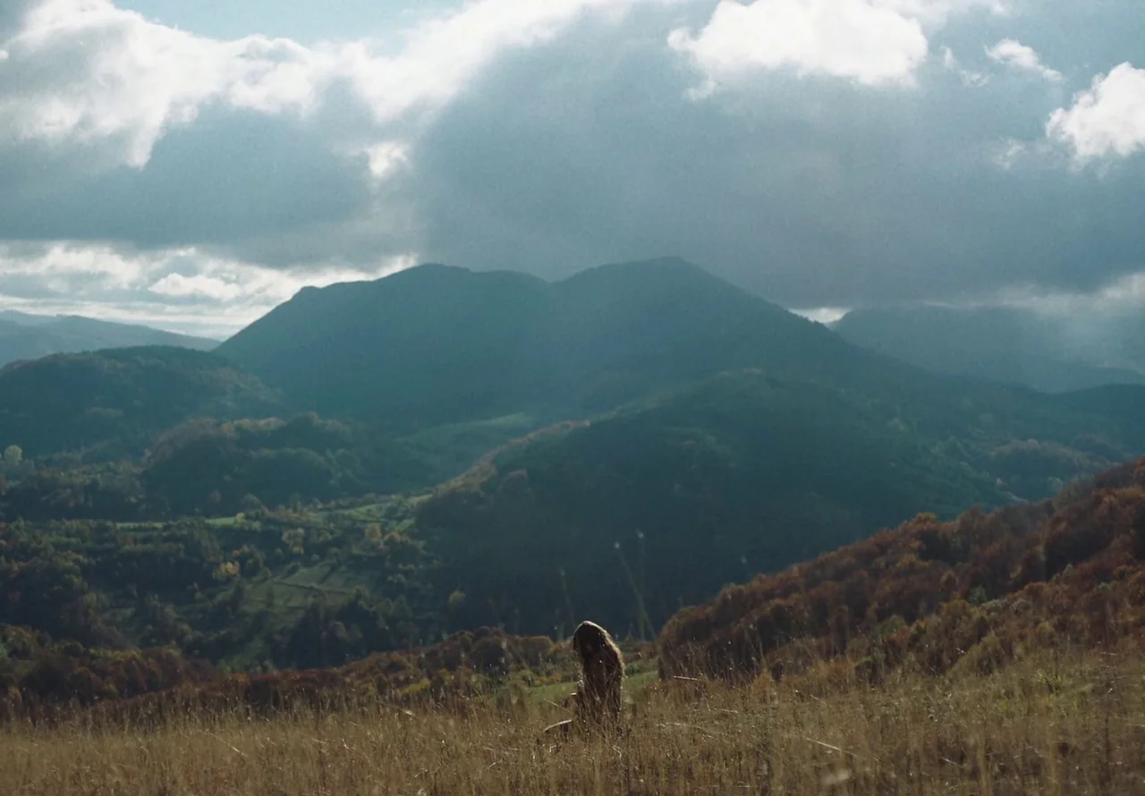 You Won’t Be Alone (2022) movie still: wide — A vast, hilly mountain landscape with dense trees and a large, cloudy sky above. A solitary person…
