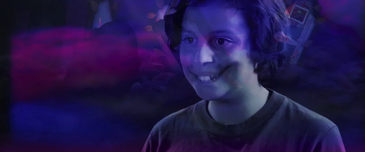 The People’s Joker (2022) movie still: medium — A teenage boy is illuminated by blue light in a dark interior environment. He faces away from the…