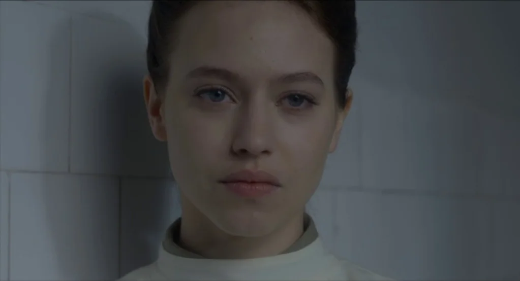 The Innocents (2016) movie still: closeup — A woman stands against a wall covered in large, pale square tiles. She looks forward with her hair…