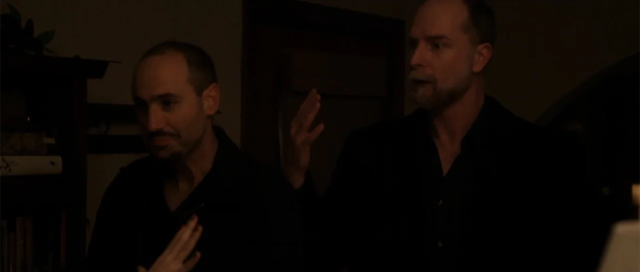 La coherencia del caos movie still: medium — A dimly lit indoor space features wooden shelves filled with books in the background. Two men engage in…