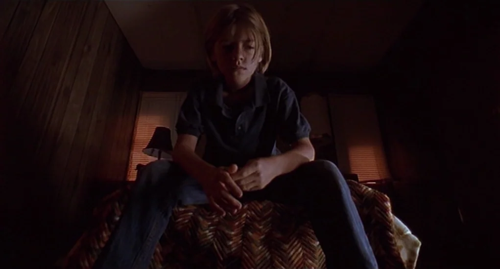 The Heart Is Deceitful Above All Things (2004) movie still: medium — A dimly lit room features wood paneling and a patterned quilt on the bed. A…