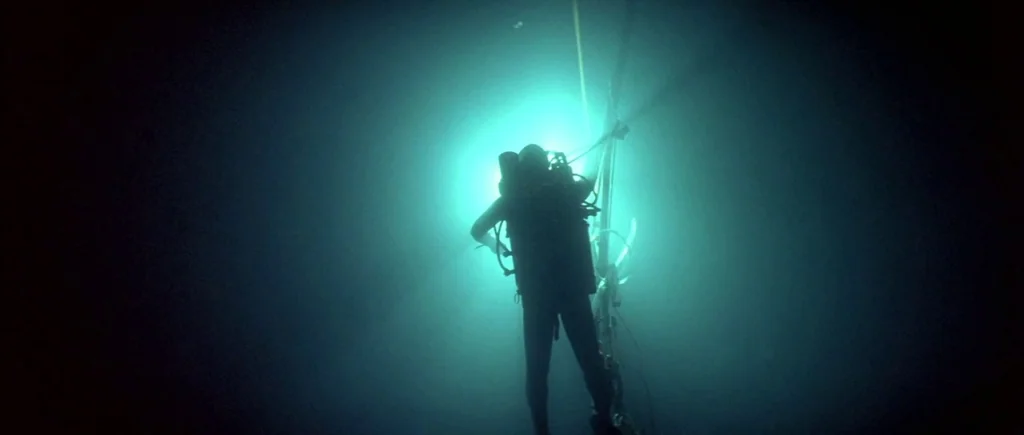 The Big Blue (1988) movie still: fullbody — An underwater scene with a lone diver framed against a bright artificial light source. The diver holds…