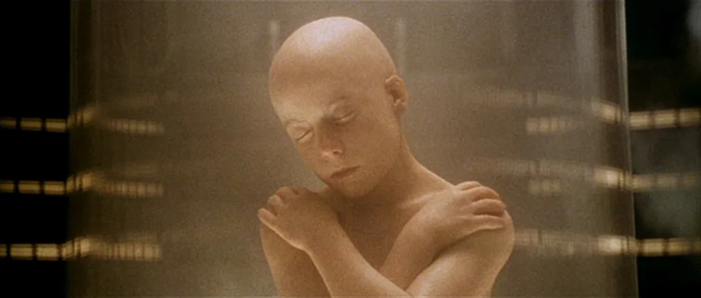 Alien: Resurrection (1997) movie still: medium — A bald child floats within a clear, cylindrical glass enclosure inside a clinical facility. The…