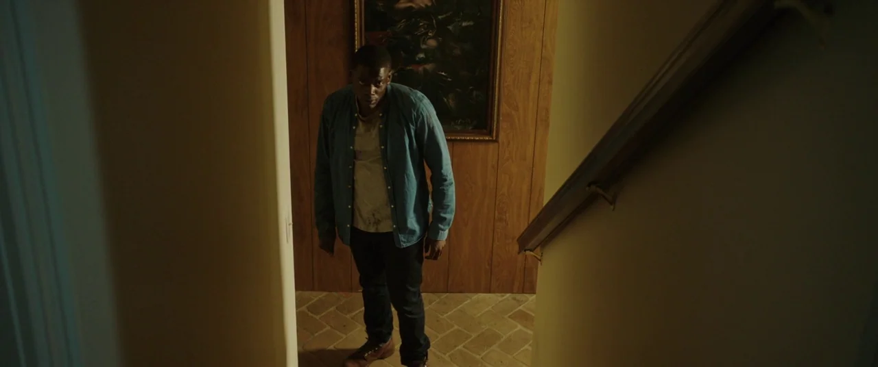 Get Out (2017) movie still: fullbody — A dimly lit indoor landing features faux wood paneling and a dark framed painting. A man stands on a tiled…