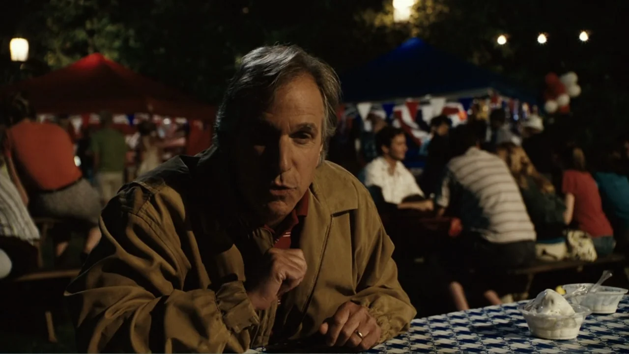 Click (2006) movie still: medium — An outdoor festival at night with picnic tables covered in blue and white checkered cloths. A man sits at a table…