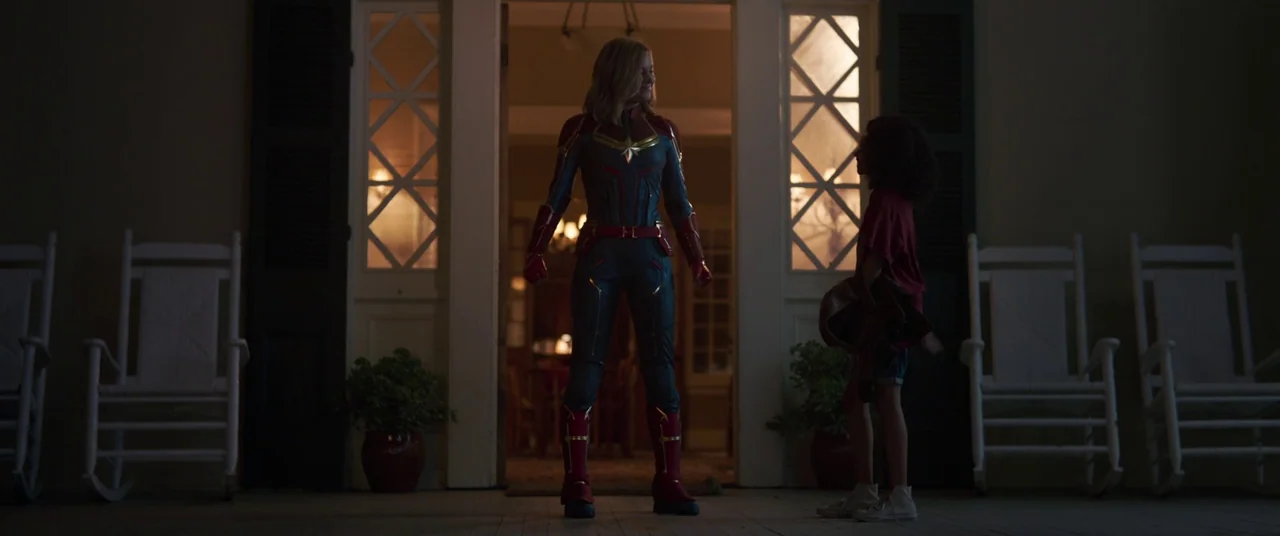 Captain Marvel (2019) movie still: fullbody — A residential porch at night with four white wooden rocking chairs. A woman in a superhero suit stands…