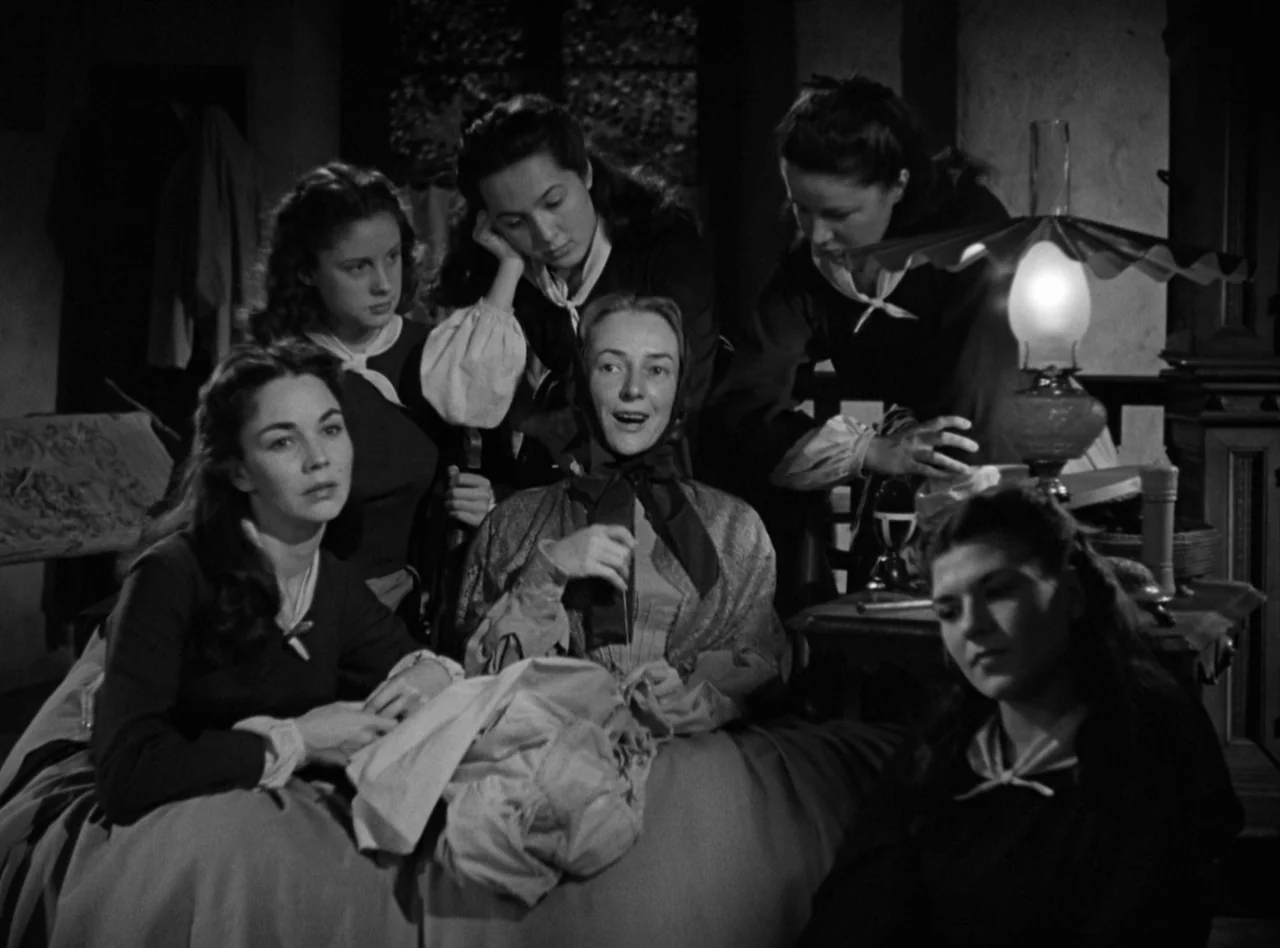 Madame Bovary (1949) movie still: medium — A group of six women in vintage attire gather around an oil lamp. One woman speaks while the others…