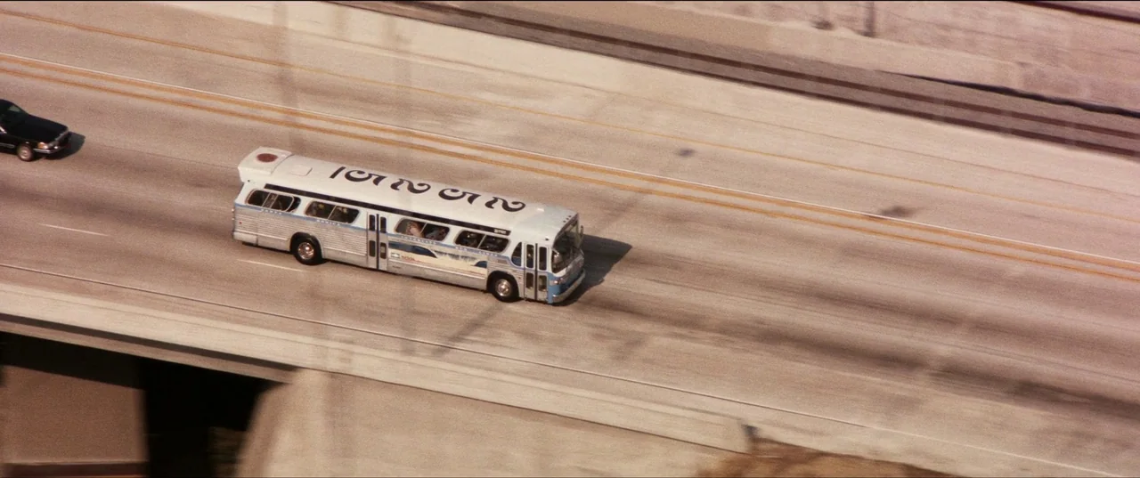 Speed (1994) movie still: wide — A wide aerial view of a multi-lane concrete freeway during daylight hours. A single bus travels along the road…