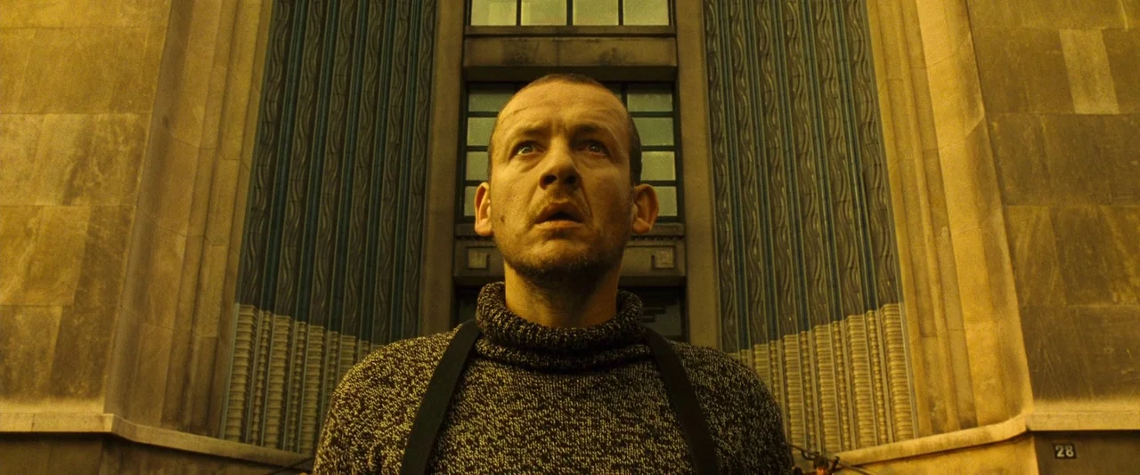 Micmacs (2009) movie still: medium — An Art Deco stone building facade rises behind a man standing in the foreground. The man looks up with a…