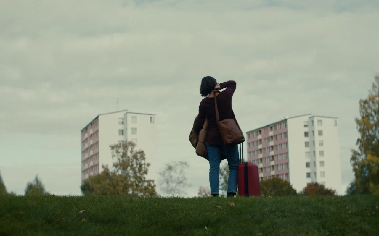 Knocking (2021) movie still: fullbody — A grassy hill in an urban area features two tall, pale apartment blocks behind. A woman holding a red…