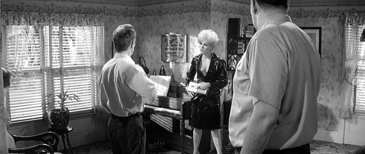 Kiss Me, Stupid (1964) movie still: fullbody — A domestic space features patterned floral wallpaper and a piano against a back wall. A woman stands…