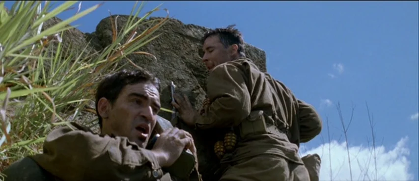 The Thin Red Line movie still: medium — Two soldiers in WWII military fatigues crouch against a large, craggy stone formation outdoors. The…