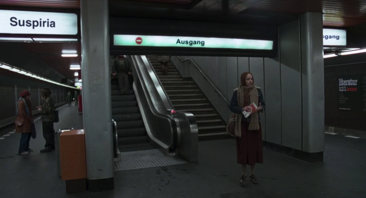 Suspiria (2018) movie still: wide — A quiet subway platform features large signage and a prominent escalator leading upward. A lone young woman with…