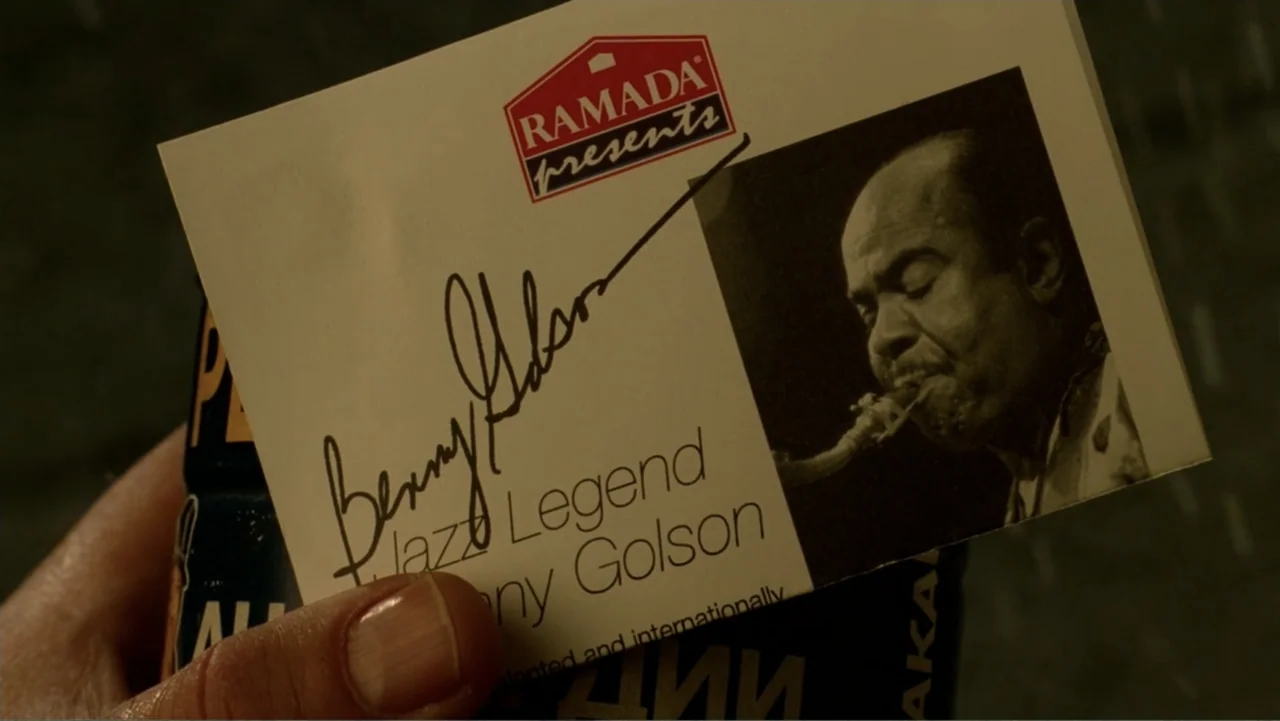 The Terminal (2004) movie still: closeup — A close-up view shows a hand holding a promotional card featuring Benny Golson. The card includes a logo…