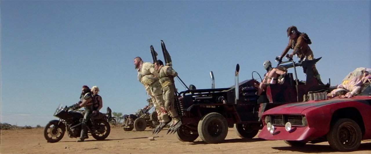 Mad Max 2 (1981) movie still: wide — A vast dry desert landscape under a clear blue sky contains several armored vehicles. A group of men in…