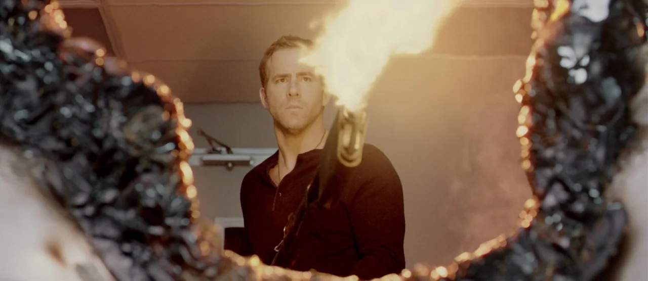 Self/less (2015) movie still: medium — A man stands in a dimly lit space behind a ragged, charred opening. He holds a flaming torch forward while…