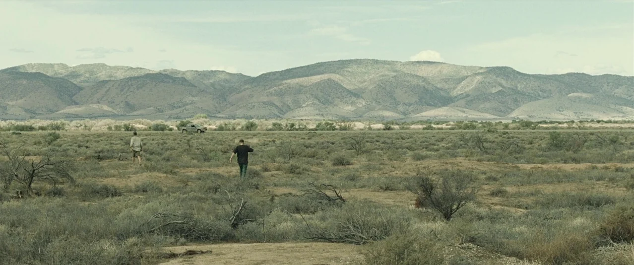 The Rover (2014) movie still: wide — A vast arid desert landscape features low scrubby brush under a large mountain range. Two men walk away from…